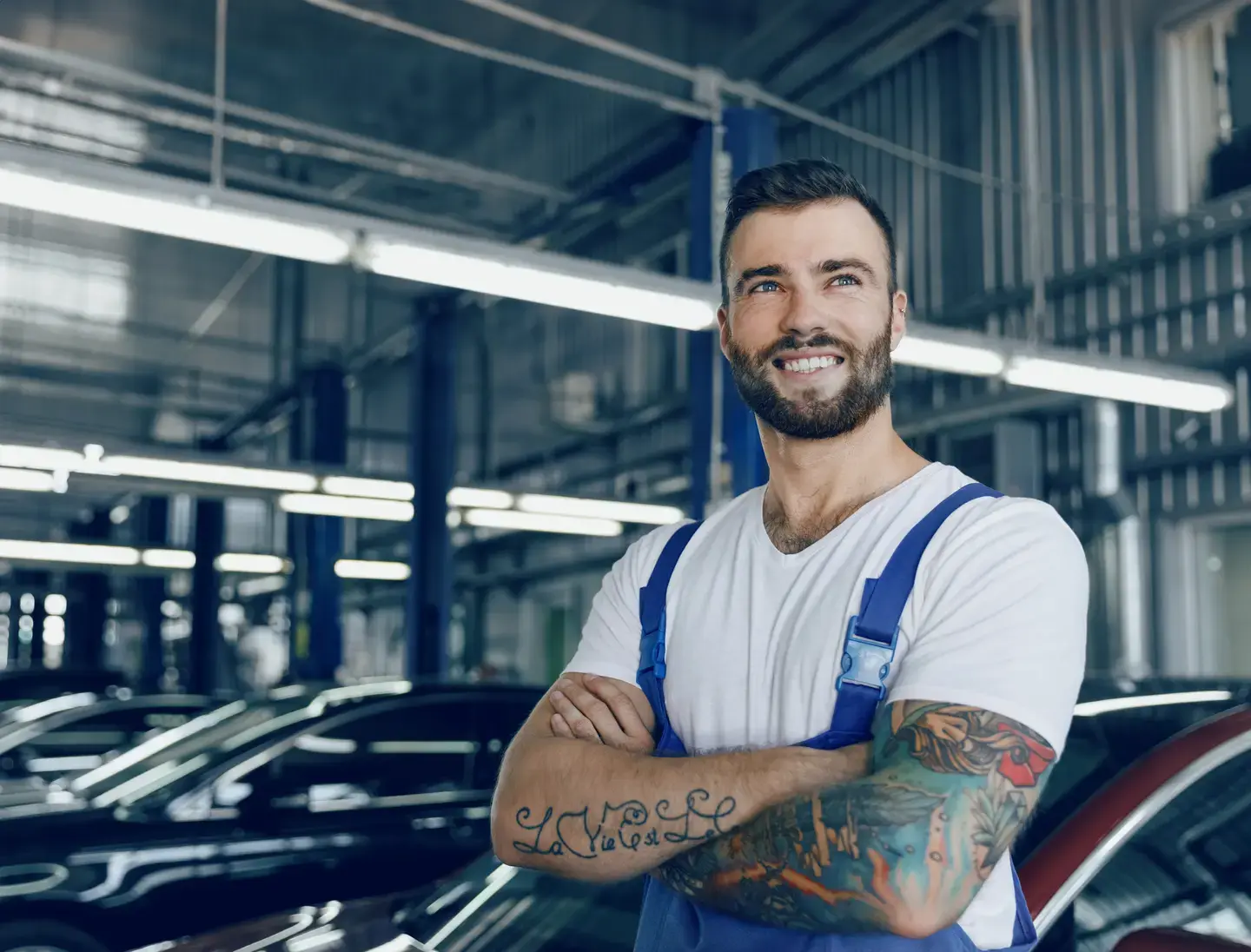 Smiling man with tattoos stands in a garage, wearing overalls.